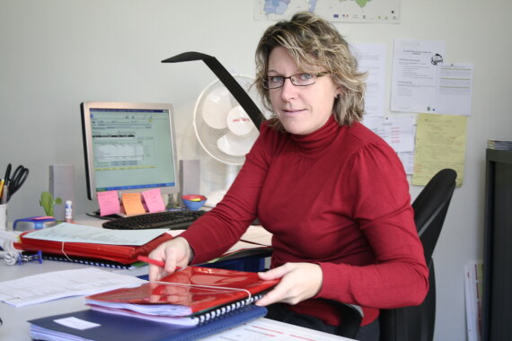 Foto von einer Mitarbeiterin von einer Behörde. Die Mitarbeiterin sitzt an einem Schreib·tisch. Die Mitarbeiterin holt 2 Mappen. Die Mitarbeiterin schaut lächelnd in die Kamera.