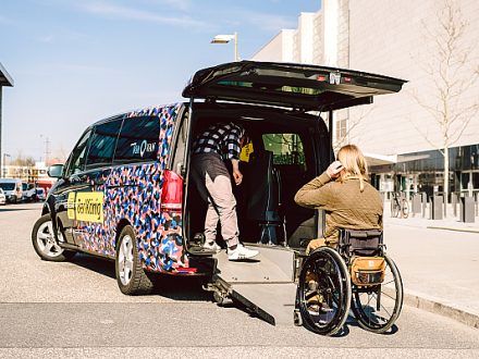 Foto: Eine Person im Rollstuhl möchte in ein Auto vom Fahr·dienst einsteigen. Die Klappe vom Koffer·raum ist offen und eine Rampe liegt auf dem Boden. Im Auto steht noch eine Person., Bild © Andi Weiland / www.gesellschaftsbilder.de