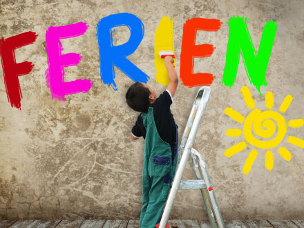 Foto von einem Jungen auf einer Leiter. Der Junge steht auf der untersten Stufe von der Leiter. Der Junge hat einen breiten Pinsel in der Hand. Der Junge malt mit dem Pinsel ein gelbes I an die Wand. An der Wand steht schon in bunten Groß·buchstaben das Wort: Ferien. Rechts unten ist eine Sonne an die Wand gemalt., Bild © S.Kobold / stock.adobe