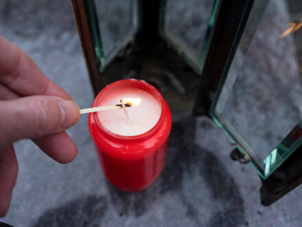 Bild © frankenphoto / stock.adobe Foto von einem roten Grab·licht. Eine Hand zündet mit einem Streich·holz die Kerze im Grab·licht an., Bild © frankenphoto / stock.adobe