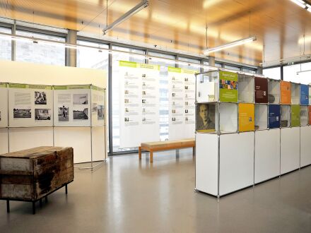 Auf dem Bild sieht man einen Teil von einer Ausstellung. In dem Raum steht ein Regal mit einem Schrank darunter. Hinter dem Regal steht eine Bank zum Sitzen aus Holz. Links neben dem Regal steht eine alte Kiste. Hinter der alten Kiste steht ein Aufsteller mit Texten an der Wand.