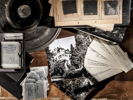Auf dem Bild sind rechts unten alte Fotos in Schwarz·weiß. Darüber sind 2 alte Ordner. Auf den Ordnern liegen ebenfalls alte Fotos. Links oben im Bild liegen alte Film·rollen. Darunter sind kleinere alte Fotos in Schwarz·weiß.