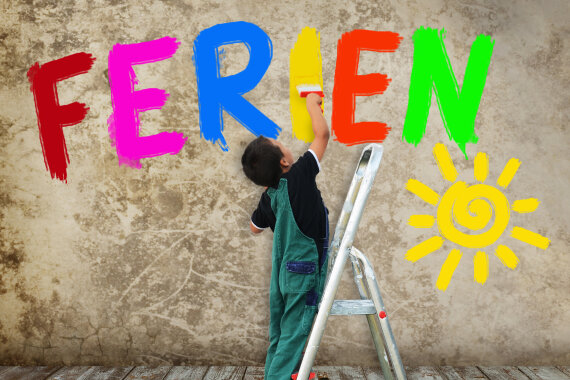 Foto von einem Jungen auf einer Leiter. Der Junge steht auf der untersten Stufe von der Leiter. Der Junge hat einen breiten Pinsel in der Hand. Der Junge malt mit dem Pinsel ein gelbes I an die Wand. An der Wand steht schon in bunten Groß·buchstaben das Wort: Ferien. Rechts unten ist eine Sonne an die Wand gemalt.