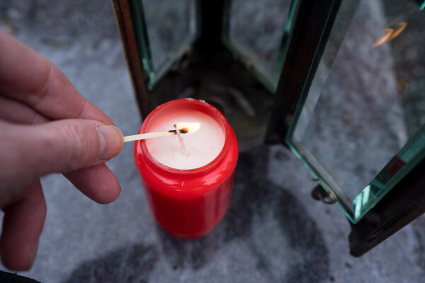 Foto von einem roten Grab·licht. Eine Hand zündet mit einem Streich·holz die Kerze im Grab·licht an.