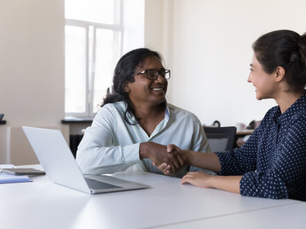 Foto von 2 Personen in einem Kurs·raum. Die beiden Personen lächeln und geben sich die Hand. Vor den Personen steht ein Laptop auf dem Tisch.