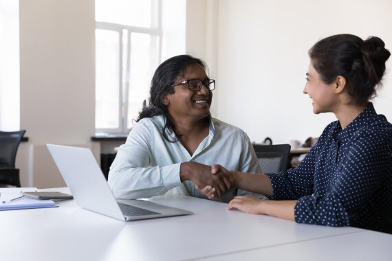 Foto von 2 Personen in einem Kurs·raum. Die beiden Personen lächeln und geben sich die Hand. Vor den Personen steht ein Laptop auf dem Tisch.