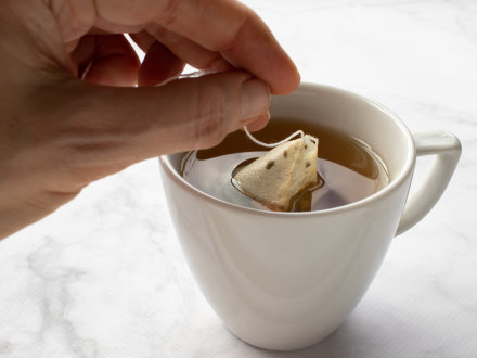 Das Bild zeigt eine Tasse Tee mit einem Tee·beutel und eine Hand.