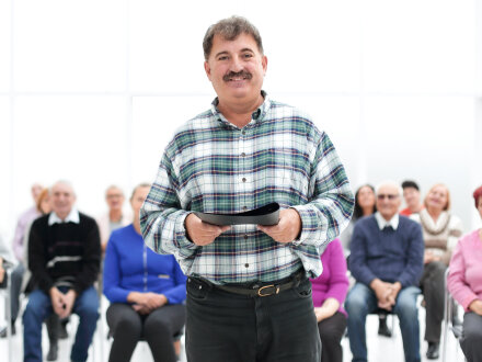 Bild © ASDF / stock.adobe Foto: Ein Mann hält einen Ordner mit Unterlagen in der Hand und lächelt. Der Mann schaut in die Kamera. Hinter ihm sitzen viele Personen und schauen freundlich., Bild © ASDF / stock.adobe