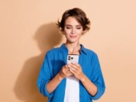 Foto: Eine Frau steht vor einer beige·farbenen Wand. Die Frau hat ein Handy in der Hand und schaut auf das Display., Bild © deagreez / stock.adobe