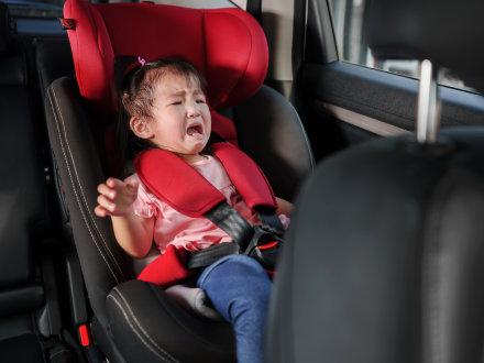 Das Bild zeigt ein kleines Mädchen in einem Kinder·sitz in einem Auto. Das Mädchen weint.