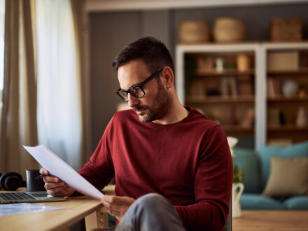 Foto von einem Mann mit einem Laptop. Der Mann sitzt am Tisch. Der Mann liest auf einem Blatt Papier.