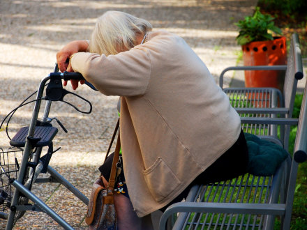 Das Bild zeigt eine ältere schwache Frau auf einer Park·bank.