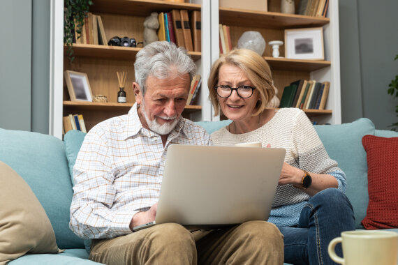 Foto von einem älteren Mann und einer älteren Frau auf einem Sofa. Der Mann hat einen Laptop auf dem Schoß. Die beiden schauen gemeinsam auf den Bildschirm.