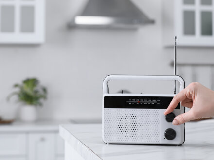 Foto von einem Radio·gerät auf einem Tisch. Eine Hand dreht an dem Knopf für die Laut·stärke., Bild © New Africa / stock.adobe