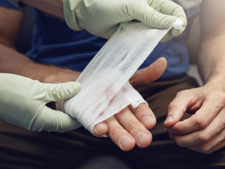Das Bild zeigt eine Person mit einer verletzten Hand. Ein Arzt wickelt um die verletzte Hand einen Verband.