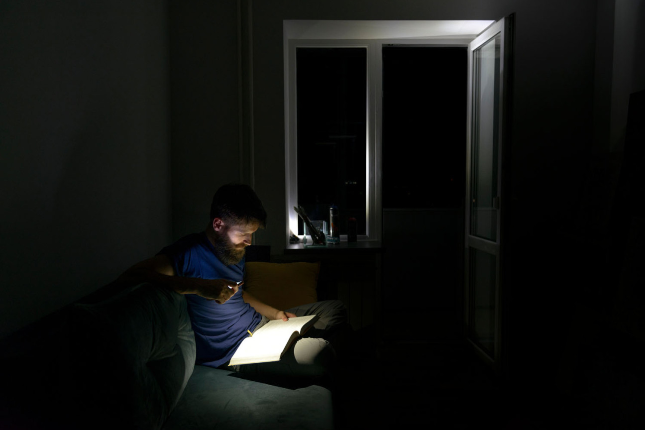 Ein Mann liest in einer Wohnung bei völliger Dunkelheit mit einer Taschenlampe ein Buch., Bild © Artur Abramiv / AdobeStock