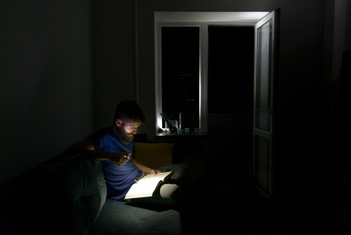 Ein Mann liest in einer Wohnung bei völliger Dunkelheit mit einer Taschenlampe ein Buch.