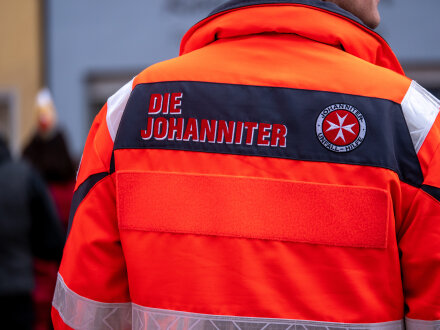 Foto von einem Mitarbeiter von der Johanniter Unfall·hilfe in einer Jacke. Auf der Jacke sieht man das Zeichen von den Johannitern und das Logo., Bild © Alex / stock.adobe