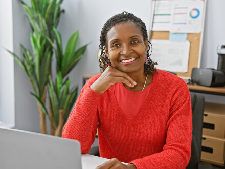 Foto von einer freundlichen Frau. Die Frau lächelt. Die Frau sitzt in einem Büro an einem Schreib·tisch. Auf dem Tisch steht ein aufgeklappter Laptop., Bild © Krakenimages.com / stock.adobe