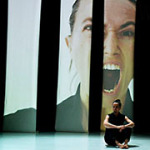Eine schreiende Tänzerin sitzt auf einem Bühnenboden. Im Hintergrund ist eine große Projektion von ihr zu sehen.