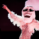 Eine Schauspielerin mit einer riesigen Totenschädel-Maske auf dem Kopf und einer rosafarbenen Federboa, breitet die Arme aus. Im Hintergrund ist das Word EXIT zu lesen.