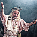 Ein Schauspieler mit Dornenkrone auf dem Kopf und einem großen Kreuz um den Hals mit nach oben gestreckten Armen.