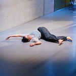 Eine Tänzerin liegt auf einer Bühne, vor einem Tisch mit einem Laptop, auf dem Boden, während das Publikum um die Bühne außen herumsteht.