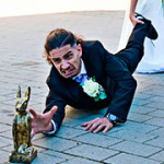 Ein junger, am Boden liegender Ehemann greift nach einer goldenen Statue, während ihn seine Braut an seinem Bein zieht und versucht wegzuschleifen