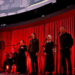 Mehrere schwarz gekleidete Personen stehen auf einer Bühne vor einem roten Vorhang