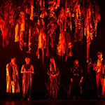 Schauspieler stehe auf einer rot beleuchteten Bühne unter Bändern, die von der Decke hängen