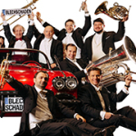 Viele Orchester-Musiker stehen und sitzen um ein rotes Cabriolet herum und halten ihre Musikinstrumente in die Höhe