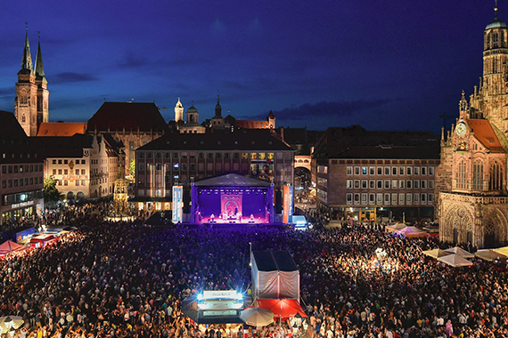 Menschenmenge vor einer beleuchteten Konzertbühne auf dem Nürnberger Hauptmarkt bei Nacht, umgeben von historischen Gebäuden und Kirchen.
