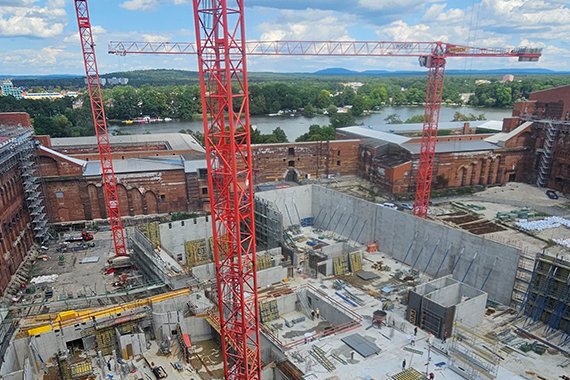 Große Baustelle mit mehreren roten Baukränen, tiefem Betonfundament und umgebenden Gerüsten. Im Hintergrund alte Backsteinmauern und dahinter ein See mit Bäumen am Ufer.