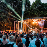 Große Menschenmenge bei einem Open-Air-Konzert vor einer beleuchteten Bühne vor Bäumen.
