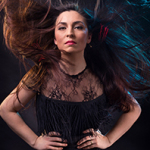 Lange, wehend aufgewirbelte Haare umrahmen ein entschlossen blickendes Gesicht. Die Frau trägt ein schwarzes Kleid mit Spitzenoberteil und Fransen, vor dunklem Hintergrund mit blauen Lichtakzenten.