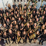 Große Musikgruppe in schwarzer Kleidung steht in einem hellen Foyer und blickt lächelnd nach oben. Viele halten Blas- oder Blechblasinstrumente wie Klarinetten, Hörner, Saxofone, Posaunen und Tuben.