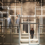 Bühnenbild mit einem Gerüst aus leuchtenden, würfelförmigen Rahmen in mehreren Ebenen. Vier Personen in grauen Anzügen stehen in unterschiedlichen Höhen, umgeben von Nebel und Lichtstrahlen.