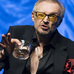 Ein Mann mit gelblicher Brille auf einer Bühne, trägt ein schwarzes Hemd mit geöffnetem Kragen und schwarzem Jackett. Er hält ein Whiskey-Glas in der Hand.