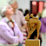 Holzskulptur einer Frau, die sich nach vorn beugt, mit einem Kind auf dem Rücken. Die Figur steht auf einem Sockel inmitten einer Ausstellung, umgeben von unscharfen Besucherinnen und Besuchern im Hintergrund.