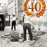 Drei Personen arbeiten auf einem gepflasterten Hof zwischen Gebäuden, umgeben von Werkzeugen, Eimern, Steinen und Erdmaterial; im Hintergrund steht ein Fahrzeug mit geöffneten Hecktüren.