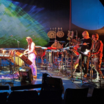 Musikerinnen und Musiker stehen auf einer Bühne mit verschiedenen Schlaginstrumenten, Keyboards und Percussion. Im Hintergrund eine großflächige, blau-grün leuchtende Projektion, darüber Scheinwerfer mit blauem Licht.