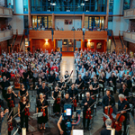 Großes Orchester mit Streich- und Blasinstrumenten vor einem applaudierenden Publikum in einer modernen, großen Halle