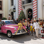 Gruppe fröhlicher Menschen in sommerlicher Kleidung und Weihnachtsmützen steht vor einem pinken Oldtimer mit goldener Schleife. Dahinter ein geschmückter Weihnachtsbaum, rot-weiß gestreifte Säulen, Schneemann und Nussknacker vor einem festlich dekorierten Gebäude.