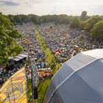 Eine große Menschenmenge sitzt dicht gedrängt auf einer Wiese zwischen Bäumen, viele mit Decken und Sonnenschirmen. Im Vordergrund Teile einer Bühne mit Technik und Überdachung, oben rechts ein Stern mit der Aufschrift „Eintritt frei“.