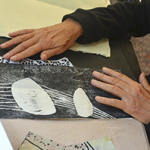 Zwei Hände bearbeiten eine schwarz-weiße Druckgrafik mit abstrakten Formen und Linien auf einem Tisch, umgeben von weiteren Papier- und Kunstwerken. Ein blauer Stern mit der Aufschrift „Eintritt frei“ ist eingeblendet.