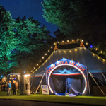 Beleuchtetes Zirkuszelt mit der Aufschrift „Comoedia Mundi“ auf einer nächtlichen Wiese, umgeben von Bäumen und kleinen Ständen. Besucherinnen und Besucher schlendern gemütlich zwischen Zelten und Imbissständen unter Lichterketten hindurch.