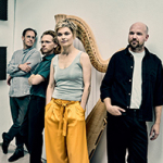 Vier Personen, im Vordergrund eine Frau mit kurzen blonden Haaren, orangfarbener Hose und grauem Trägertop. Dahinter steht eine große Konzertharfe.