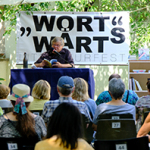 Ein Mann liest im Freien an einem Tisch mit Mikrofon und Getränken vor Publikum, hinter ihm ein Banner mit der Aufschrift „WortWärts Literaturfest“. Im Hintergrund ein Regal mit Büchern vor einem Baum.
