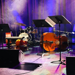 Bühne mit Jazz-Instrumenten im Scheinwerferlicht: Kontrabass, Marimbaphon, Baritonsaxophon, Schlagzeug und weitere Perkussion sind spielbereit positioniert, umgeben von Mikrofonen und Notenständern, vor einem farbig beleuchteten, nebelverhangenen Bühnenvorhang.