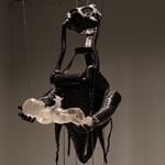 Schwarze, hängende Figur mit Gasmaske und kniehohen Stiefeln, die ein durchsichtiges, liegendes Babymodell in den Händen hält.